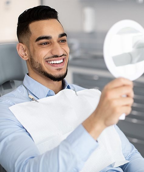 Man in blue shirt in dental chair smiling at his reflection holding a hand mirror