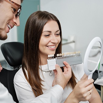 Woman with long brown hair smiling into hand mirror as dentist holds shade guide to her teeth